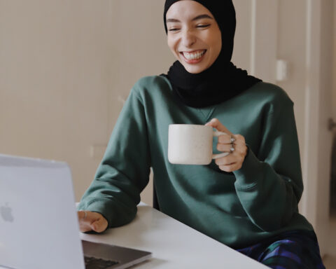 Photo of a woman in front of a laptop, wearing a sweatshirt and pajama pants, holding a mug.
