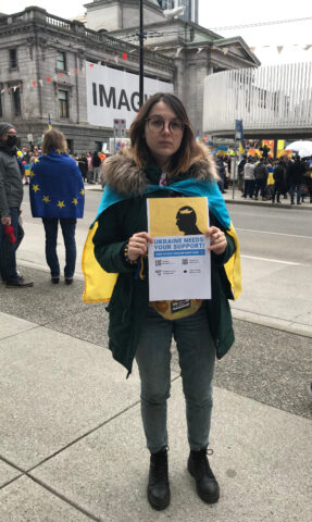 A Ukrainian Canadian woman with the Ukrainian flag draped around her shoulders holds up a sign that reads "Ukraine needs your support"