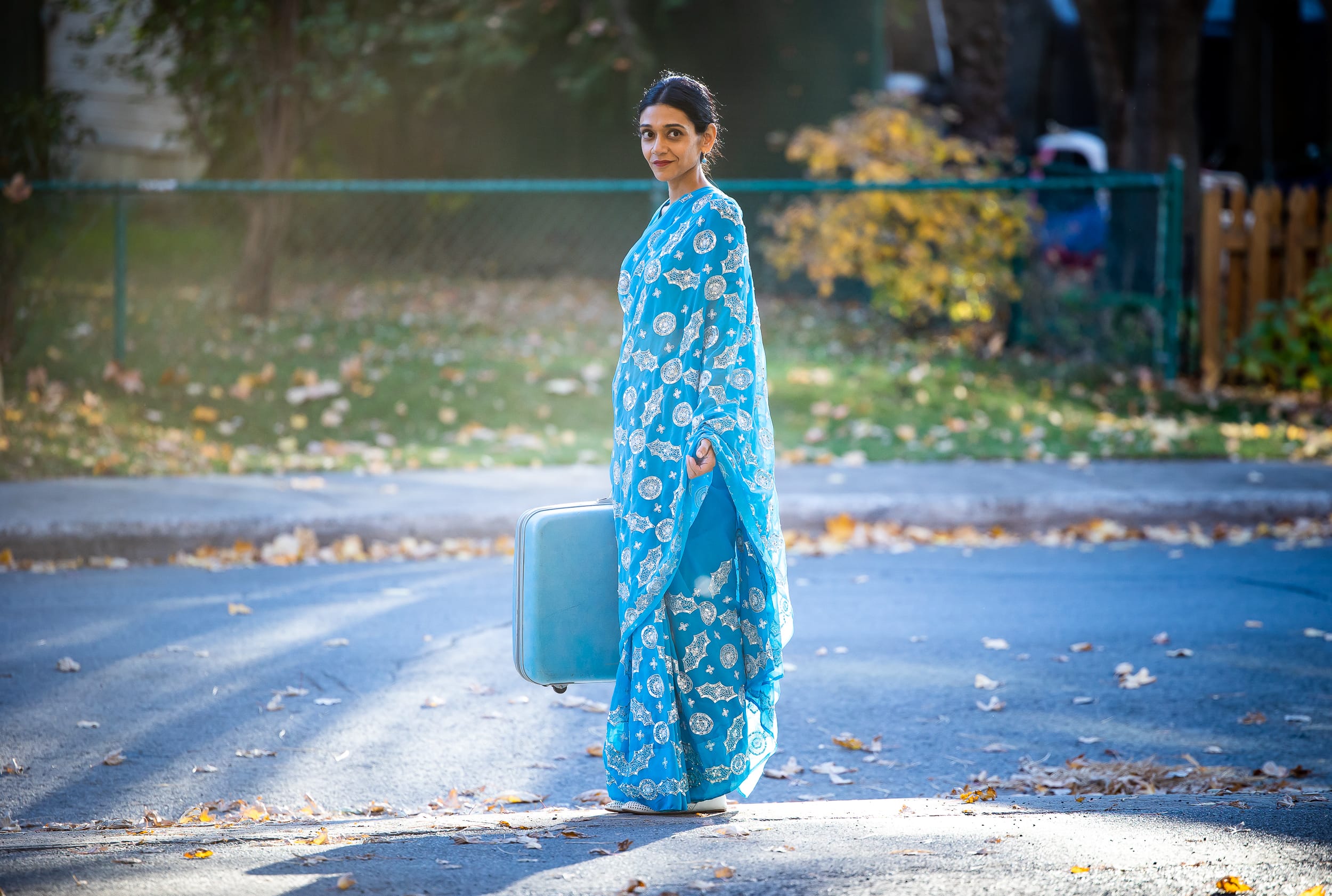 A woman in a bright blue sari stands on a residential street holding a silver suitcase in a still from the film Calorie.