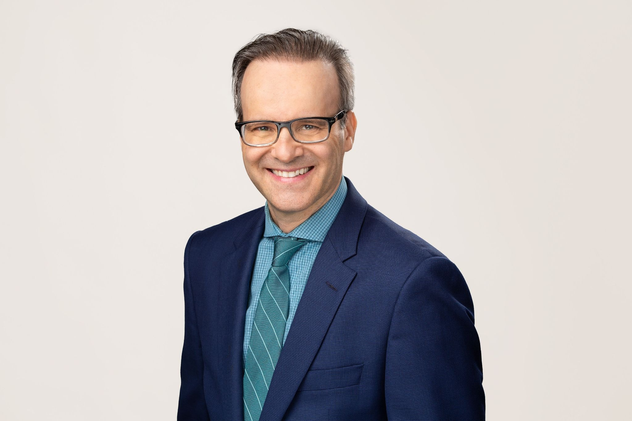 Portrait of immigration lawyer Mario Bellissimo wearing glasses, a navy suit, teal shirt and striped tie against a light background.
