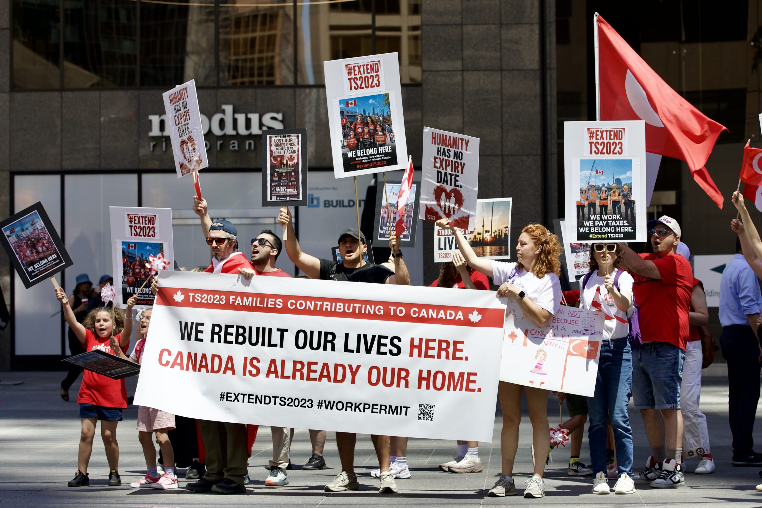 Adults and children march in downtown Toronto holding Turkish and Canadian flags, a large “We rebuilt our lives here” banner and signs calling for the extension of TS2023 work permits.