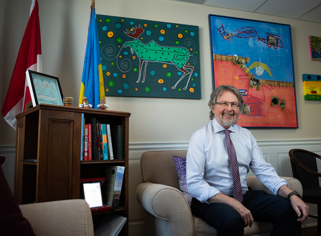 A man in a dress shirt and tie sits smiling in an office beside Canadian and Ukrainian flags, with colourful artwork displayed on the wall behind him.