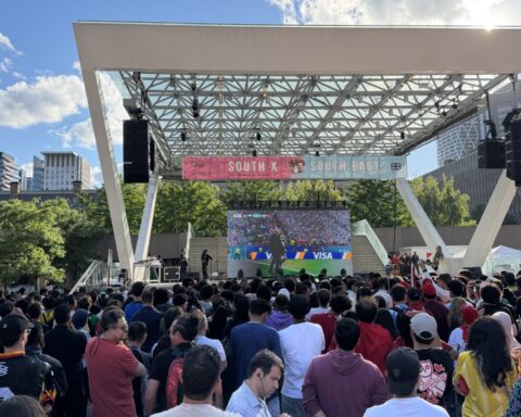 A large crowd gathers under an outdoor canopy at Nathan Phillips Square in Toronto, watching a World Cup match on a big screen, with the city skyline visible in the background.