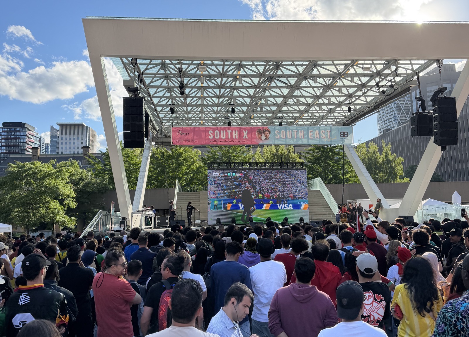 A large crowd gathers under an outdoor canopy at Nathan Phillips Square in Toronto, watching a World Cup match on a big screen, with the city skyline visible in the background.