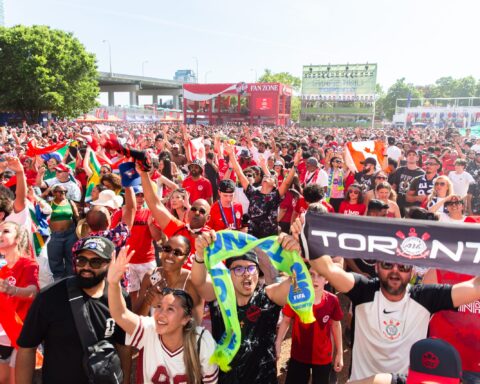 A dense crowd of soccer fans packs the FIFA Fan Festival grounds in Toronto, most wearing red Canada jerseys and waving Canadian flags, with several also holding South African flags. In the foreground, fans cheer, wave scarves, and hold up a black and white Toronto-branded banner. In the background, a Coca-Cola Fan Zone structure and a large "FanFestival Toronto" screen rise above the crowd, framed by trees and a highway overpass.