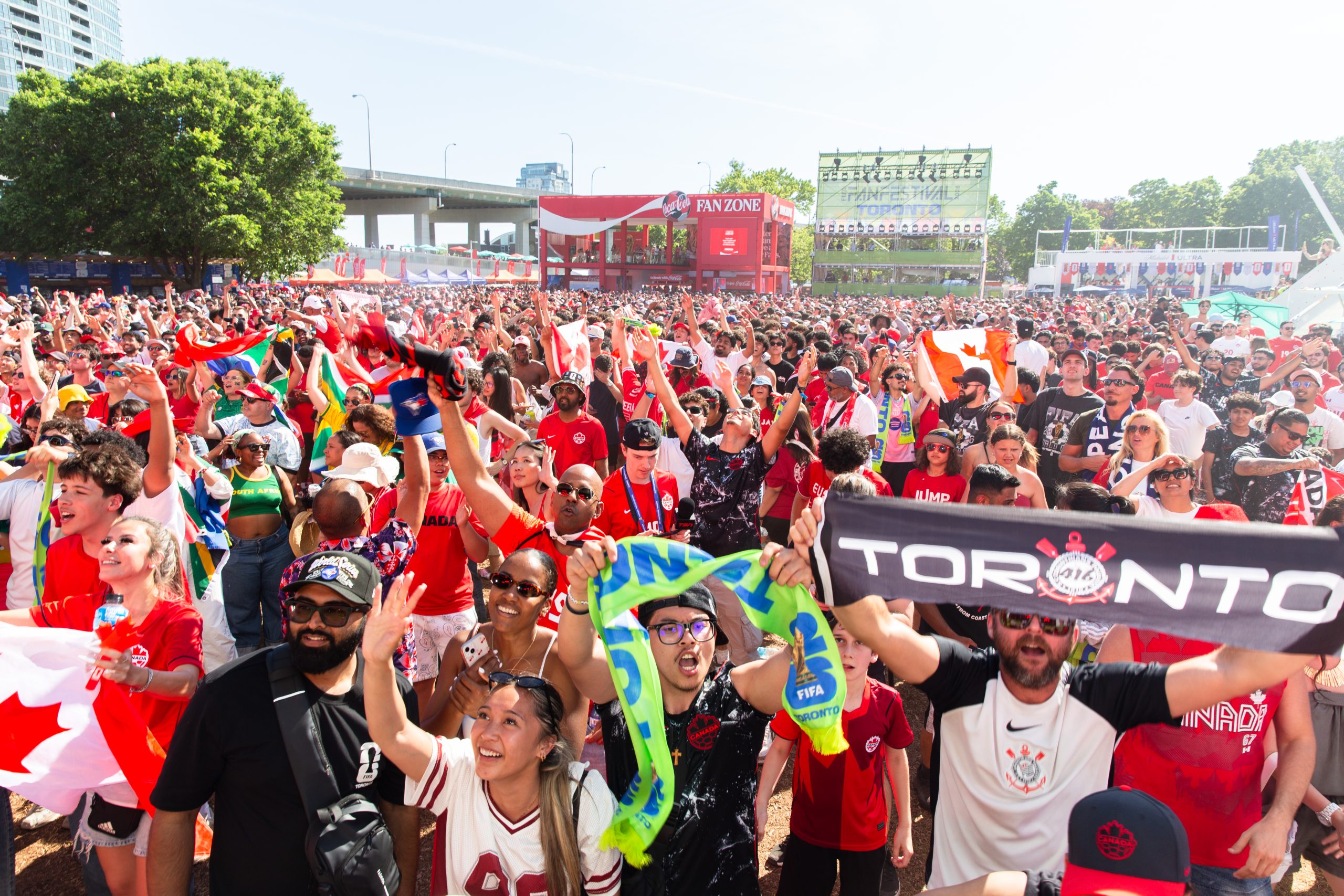 A dense crowd of soccer fans packs the FIFA Fan Festival grounds in Toronto, most wearing red Canada jerseys and waving Canadian flags, with several also holding South African flags. In the foreground, fans cheer, wave scarves, and hold up a black and white Toronto-branded banner. In the background, a Coca-Cola Fan Zone structure and a large "FanFestival Toronto" screen rise above the crowd, framed by trees and a highway overpass.