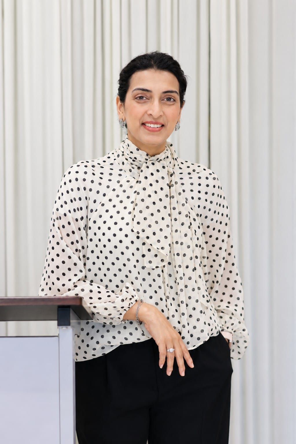 Gul Rizvi stands indoors wearing a white polka-dot blouse and black pants.