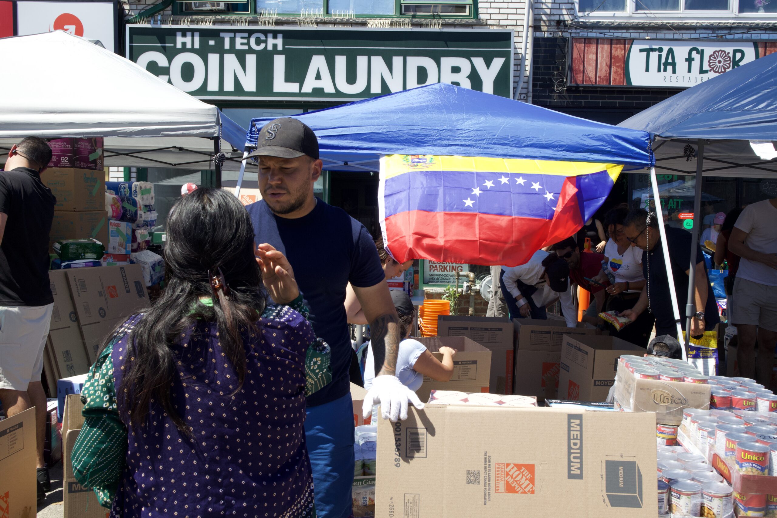 Volunteers sort boxes of donated food and supplies under tents outside Tia Flor restaurant in Toronto, with a Venezuelan flag hanging from one canopy.