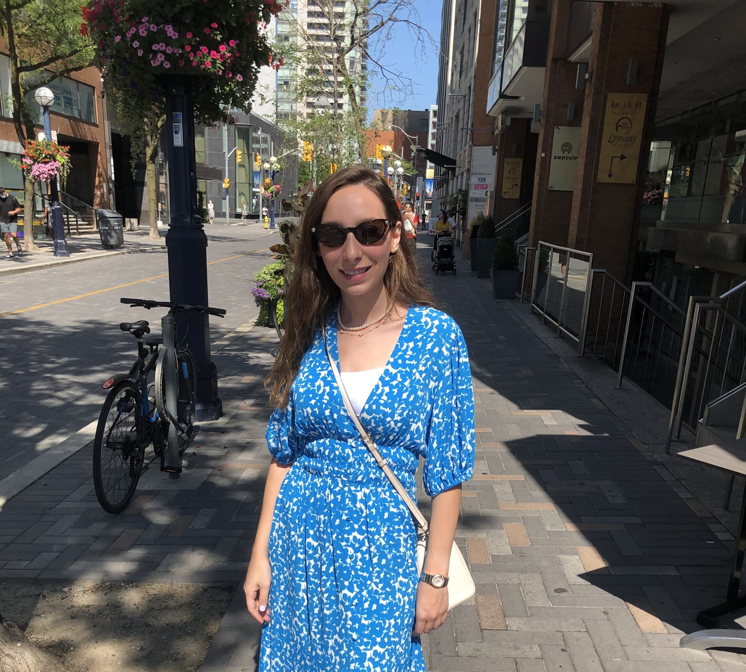 Razan Abu-Ghoush stands on a sunny downtown Toronto sidewalk, wearing a blue-and-white patterned dress and sunglasses, smiling at the camera.