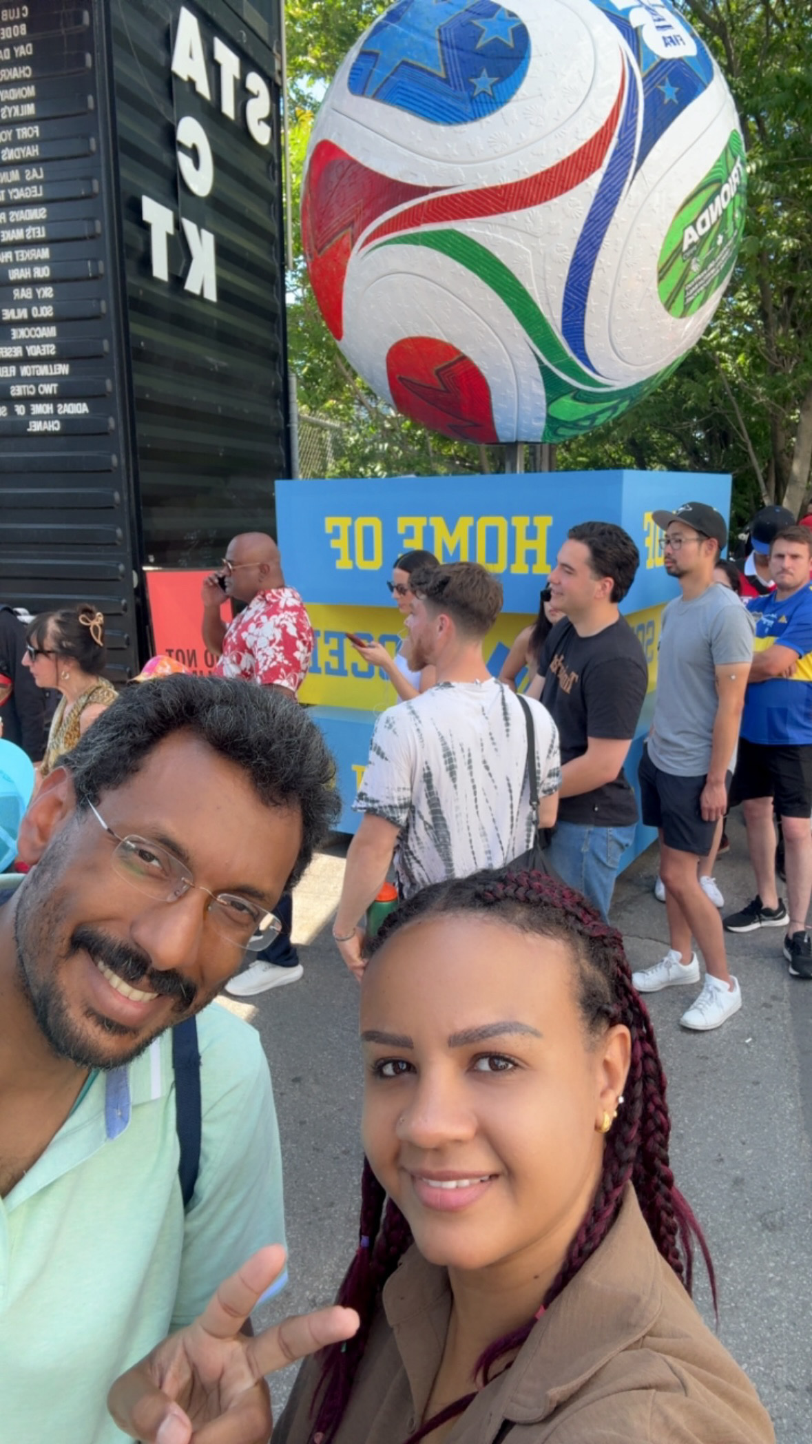 A man and a woman smile for a selfie at a World Cup fan zone in Toronto, with a giant decorated soccer ball sculpture mounted on a blue 'Home of Soccer' display behind them and a crowd of fans milling around.