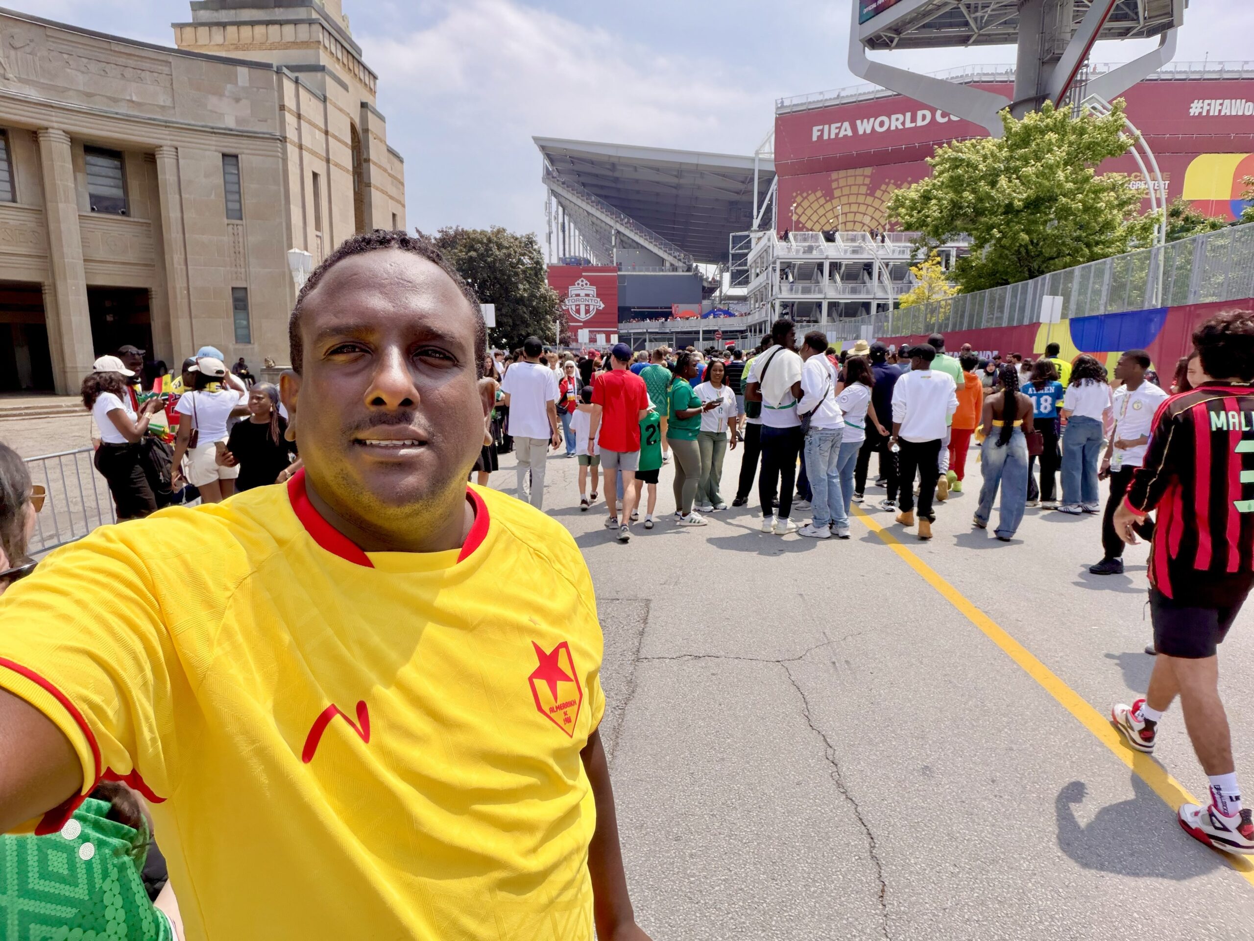 Mustafa Hussein takes a selfie outside a World Cup stadium in Toronto, wearing a yellow and red jersey, with crowds of fans and the stadium's grandstand visible behind him.