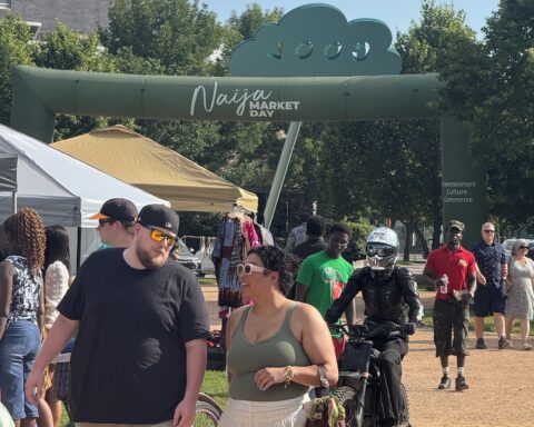 People walk past the entrance arch for Naija Market Day at an outdoor market, with vendor tents and shoppers visible on a sunny day.