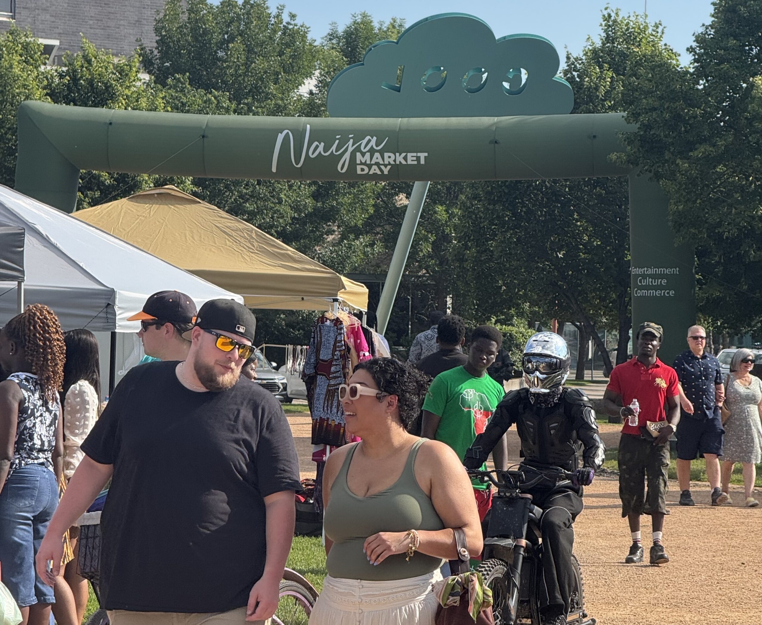 People walk past the entrance arch for Naija Market Day at an outdoor market, with vendor tents and shoppers visible on a sunny day.