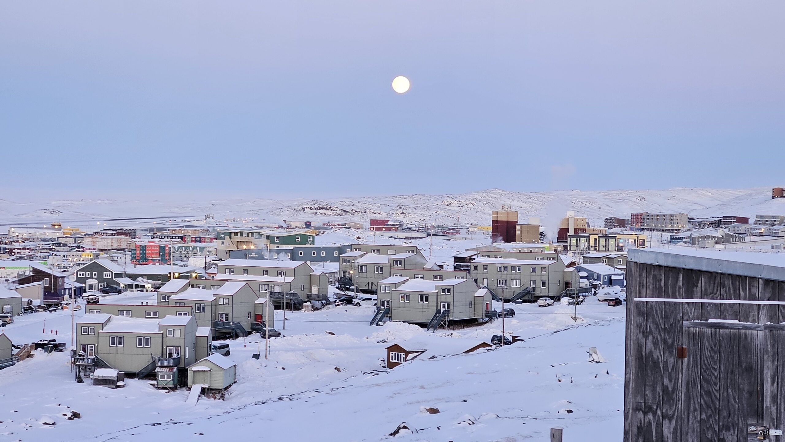 A full moon rises over rows of snow-covered houses in Iqaluit, Nunavut, with snow-capped hills in the background.