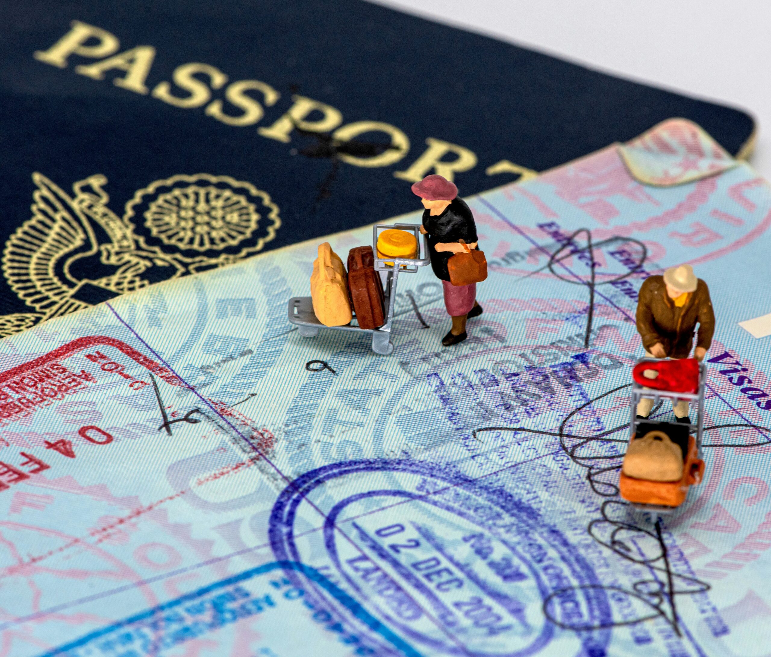 Miniature figurines pushing luggage carts across a passport page covered in visa stamps.
