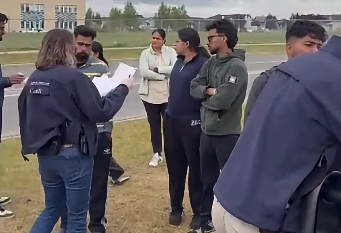 A uniformed officer holds a clipboard while speaking to a group of students standing in a semicircle on a grassy field, with an apartment building and street visible in the background.