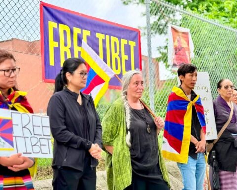 A group of Tibetan demonstrators stand along a chain-link fence in Ottawa, several wrapped in the Tibetan flag and dressed in black.