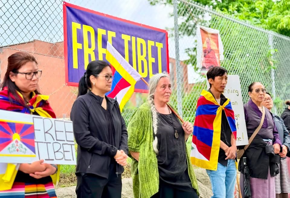 A group of Tibetan demonstrators stand along a chain-link fence in Ottawa, several wrapped in the Tibetan flag and dressed in black.