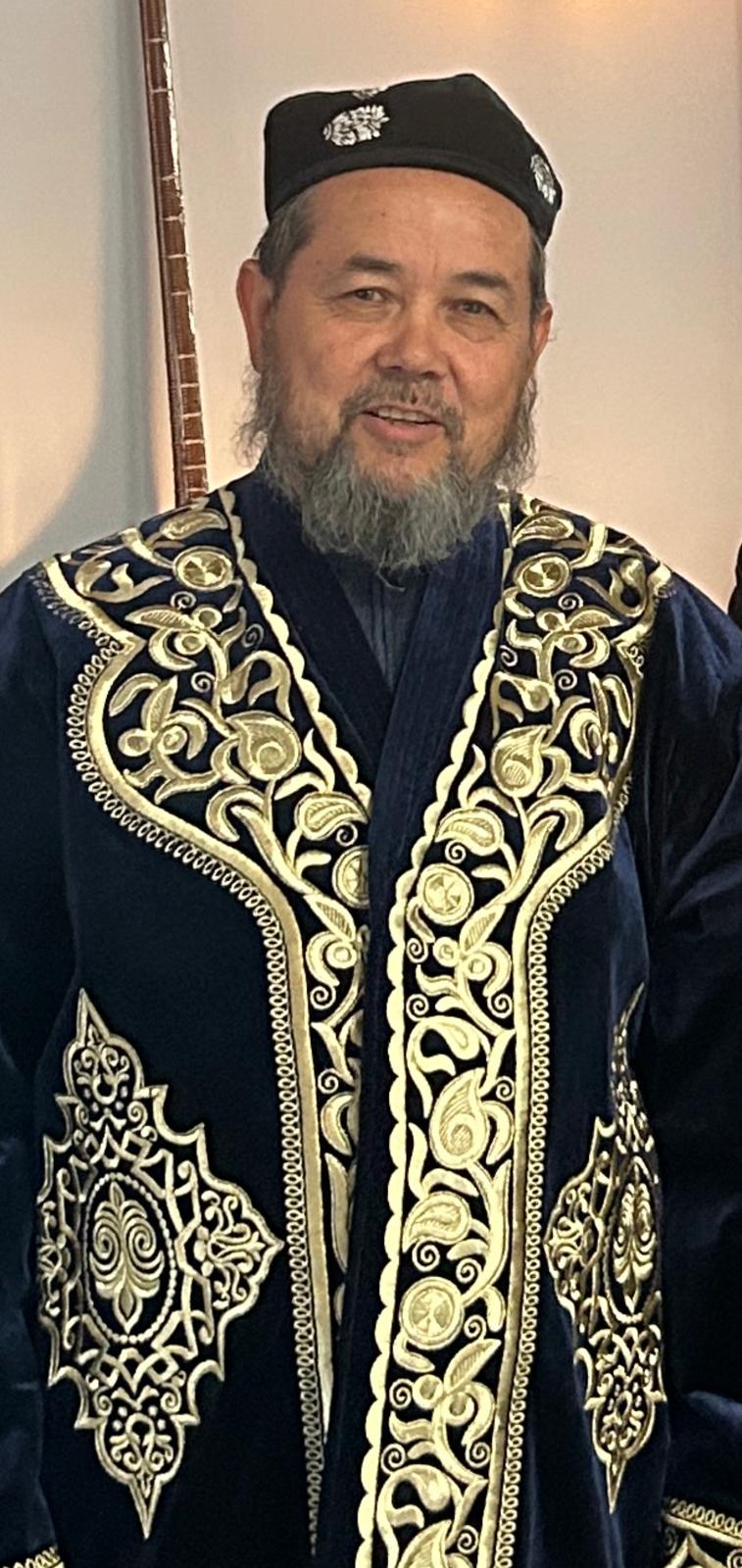 Abdulahat Nur, wearing a traditional dark blue and gold embroidered Uyghur robe and cap, stands indoors against a light-coloured wall.