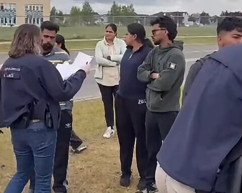 A uniformed officer holds a clipboard while speaking to a group of students standing in a semicircle on a grassy field, with an apartment building and street visible in the background